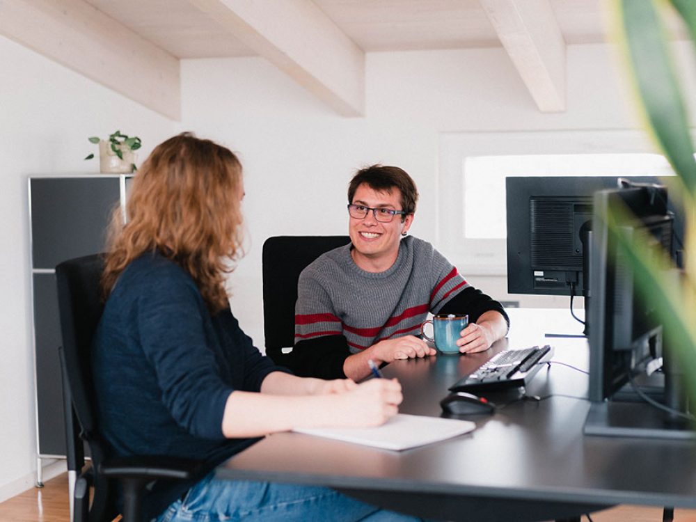 Zwei Kollegen sitzen an einem Schreibtisch und unterhalten sich freundlich.