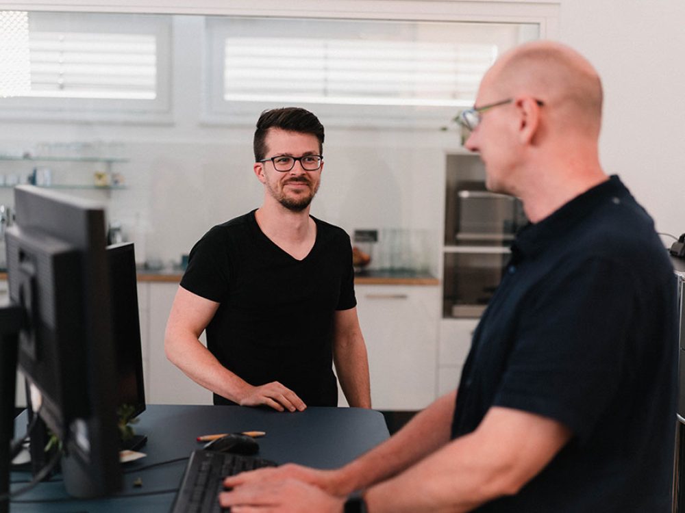 Zwei Männer unterhalten sich an einem Schreibtisch in einem Büro.